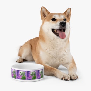May include: White ceramic dog bowl featuring a purple and green marijuana leaf design. A Shiba Inu dog is sitting beside the bowl.