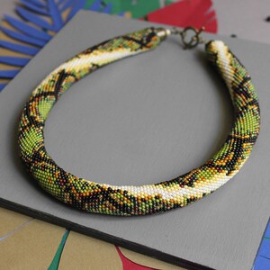 Puede incluir: Un collar de cuentas verde, blanco y negro con un patrón de piel de serpiente. El collar está sobre una superficie gris.