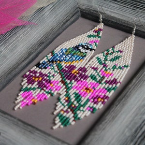 Peut inclure: Une paire de longues boucles d'oreilles en perles avec un motif floral et un oiseau. Les boucles d'oreilles sont faites de perles blanches, roses, vertes, violettes et bleues. Les boucles d'oreilles sont suspendues à des crochets argentés.