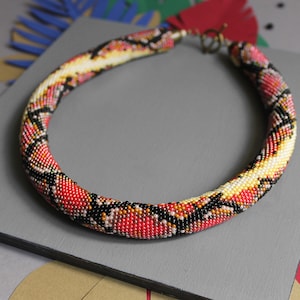 Op de afbeelding: Een rode, zwarte en gele kralenketting met een slangenhuidpatroon. De ketting is gemaakt van kleine kralen en is ongeveer 45 cm lang.