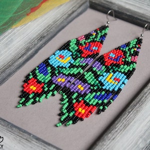 May include: Handmade beaded earrings with a floral design. The earrings feature a black background with colourful flowers in red, blue, purple, and green. The earrings are approximately 10 cm long.