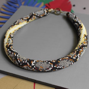Puede incluir: Un collar de cuentas con un patrón de piel de serpiente en negro, blanco, gris y marrón. El collar está hecho de cuentas pequeñas y tiene un cierre.