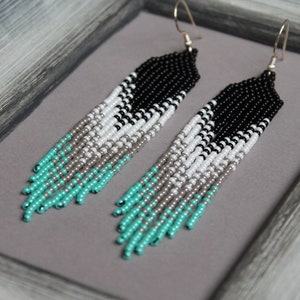 May include: A pair of long, beaded earrings with a black, white, silver, and turquoise colour scheme. The earrings are made of small, round beads and have a fringe-like design.