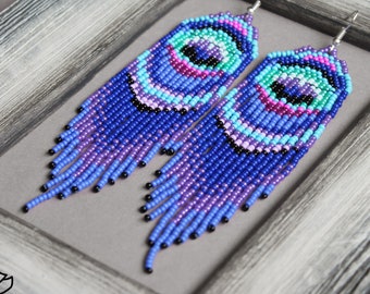 Può includere: Un paio di lunghi orecchini a perline con un design colorato a forma di occhio in tonalità di blu, viola, rosa e verde. Gli orecchini sono realizzati con piccole perline rotonde e hanno una parte inferiore a forma di frangia.