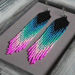 Puede incluir: Un par de pendientes largos de cuentas con un degradado de cuentas rosas, verdes, azules y negras. Los pendientes cuelgan de ganchos plateados.