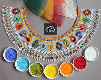 May include: A white beaded necklace with colourful geometric patterns and a fringe. The necklace is displayed on a grey surface with small bowls of beads in various colours.