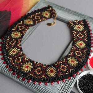 May include: A striking beaded necklace featuring a geometric design in black, red, and gold. The necklace is presented on a grey picture frame. Small bowls of gold, red, and black beads are nearby. The necklace has a gold clasp.