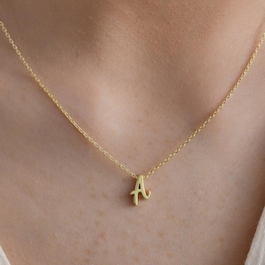 Può includere: Una delicata collana d'oro con un ciondolo a forma di lettera "A" in corsivo. Il ciondolo è appeso a una sottile catena d'oro, creando un look minimalista ed elegante. La collana è mostrata su uno sfondo morbido e neutro.