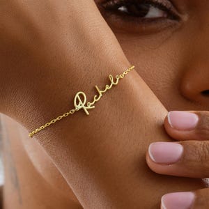 May include: A gold-colored bracelet with the name "Rachel" in cursive script. The bracelet is delicate and appears to be made of a thin chain. The image is a close-up, showcasing the bracelet on a wrist.