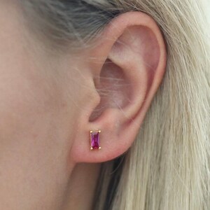 May include: A pair of gold stud earrings with a small, rectangular, pink gemstone.
