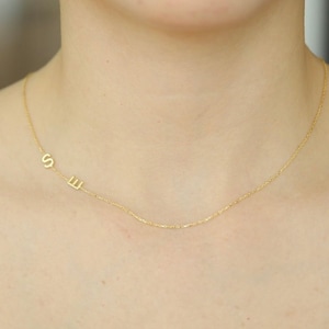 May include: A gold chain necklace with two small letters, "S" and "E", hanging from it. The necklace is worn by a person with red hair.