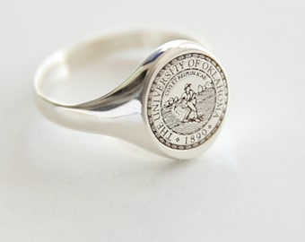 May include: A silver ring with a round seal featuring the University of Oklahoma logo and the text "The University of Oklahoma 1890".