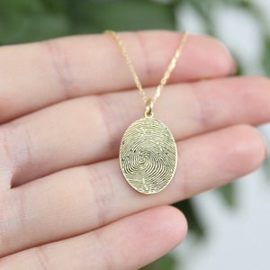 K&ouml;nnte beinhalten: Eine goldene Halskette mit einem ovalen Anh&auml;nger, der ein detailliertes Fingerabdruckdesign zeigt.