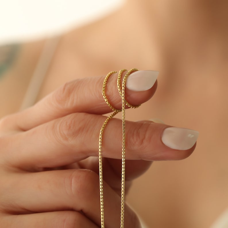 May include: A gold chain necklace with a box chain design. The chain is held in a person's hand.
