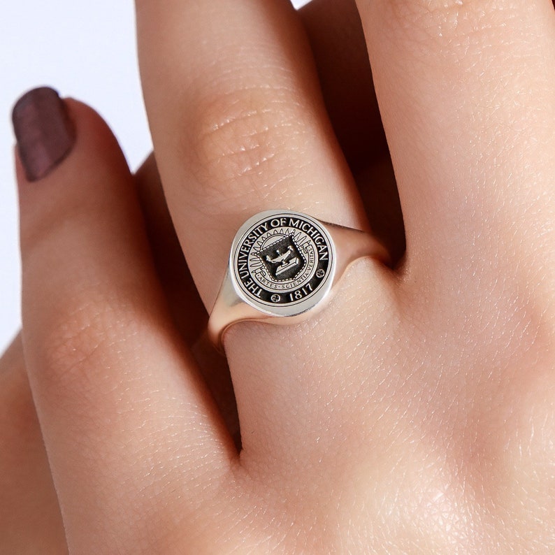 May include: A silver signet ring with a black enamel seal featuring the University of Michigan logo and the text "The University of Michigan 1817".