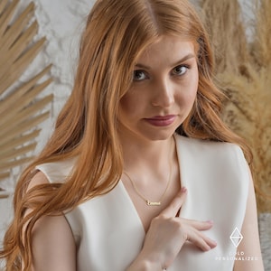 May include: A woman with long, wavy, red hair is wearing a white sleeveless top and a gold necklace with the name "Emma" on it.