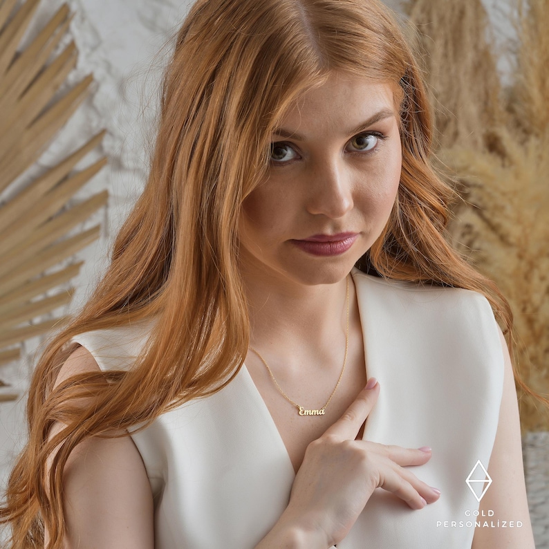 May include: A woman with long, wavy, red hair is wearing a white sleeveless top and a gold necklace with the name "Emma" on it.
