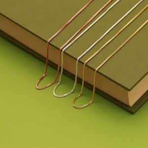 May include: Five different chain necklaces in various metal finishes, including silver, gold, and rose gold, are displayed on a green surface. The chains are arranged in a row, with the rose gold chain at the top and the silver chain at the bottom.