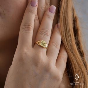 May include: A gold signet ring with a square top featuring a monogram. The ring is worn on a person's finger.