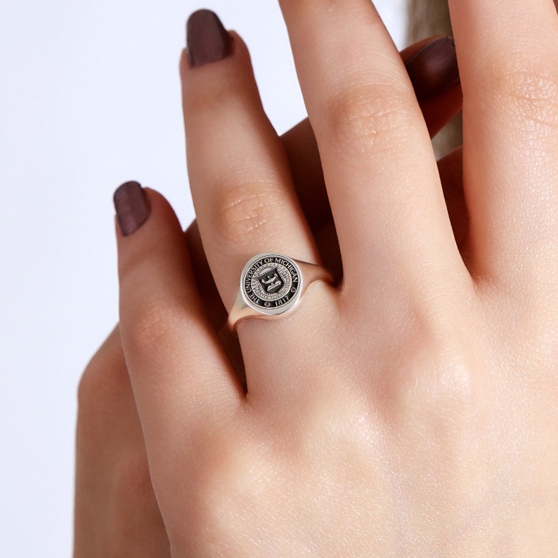 May include: A silver signet ring with a black enamel University of Michigan crest. The crest features a shield with a winged griffin and the text "University of Michigan 1817".