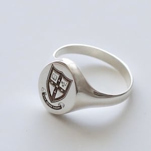 May include: A silver signet ring with a crest featuring a shield with a cross and a book, surrounded by a scroll with the text "Fide Quam Videri".