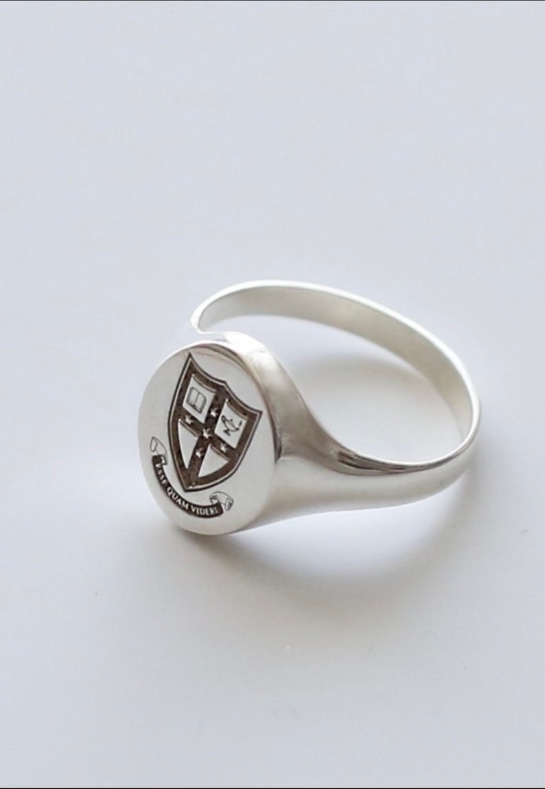 May include: A silver signet ring with a crest featuring a shield with a cross and a book, surrounded by a scroll with the text "Fide Quam Videri".