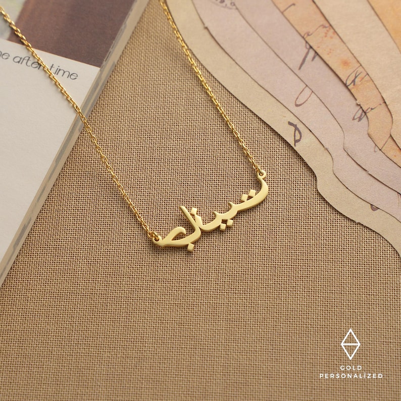 May include: A gold necklace with a personalized nameplate featuring the Arabic word "سُبْحَانَ الله" in a simple, elegant font.