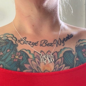 May include: A silver necklace with a monogram pendant featuring the letters "HJB". The necklace is worn by a person with a tattoo on their chest that reads "Żyć Bez Wyroku".