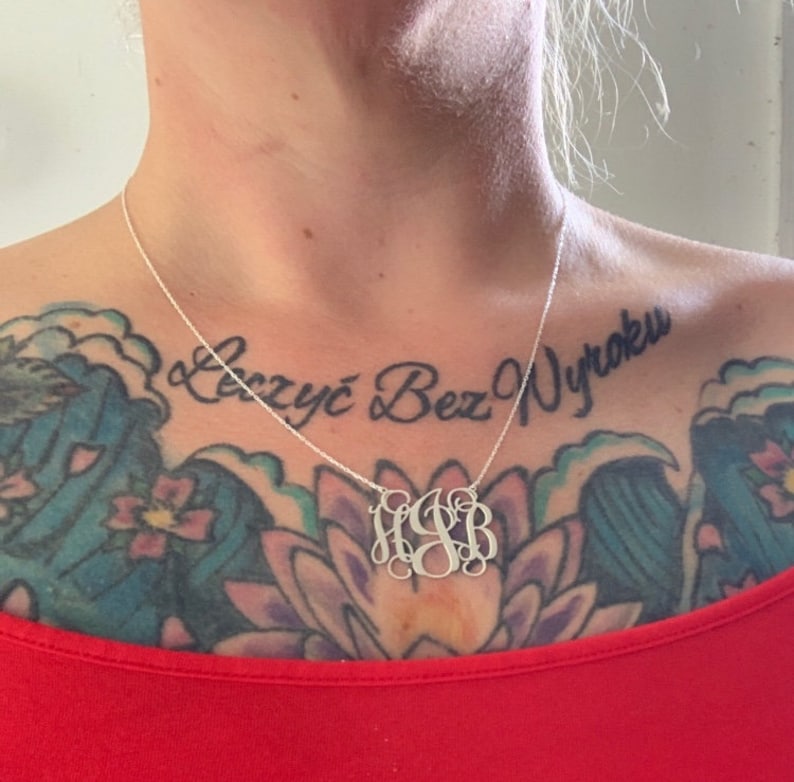 May include: A silver necklace with a monogram pendant featuring the letters "HJB". The necklace is worn by a person with a tattoo on their chest that reads "Żyć Bez Wyroku".