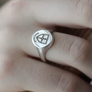 May include: A silver signet ring with a crest engraved on the face. The crest features a shield with a cross and other symbols.