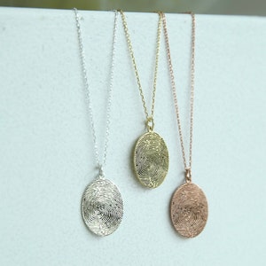K&ouml;nnte beinhalten: Drei silberne Halsketten mit ovalen Anh&auml;ngern, die ein Fingerabdruckdesign aufweisen. Die Anh&auml;nger sind in Silber, Gold und Ros&eacute;gold.
