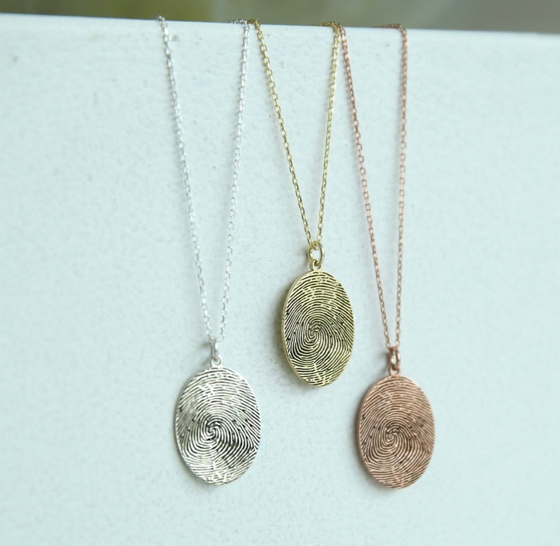 K&ouml;nnte beinhalten: Drei silberne Halsketten mit ovalen Anh&auml;ngern, die ein Fingerabdruckdesign aufweisen. Die Anh&auml;nger sind in Silber, Gold und Ros&eacute;gold.