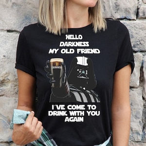 May include: Two black and white t-shirts featuring a Star Wars character, Darth Vader, holding a bottle of alcohol. The text on the shirts reads "Hello darkness my old friend, I've come to drink with you again."