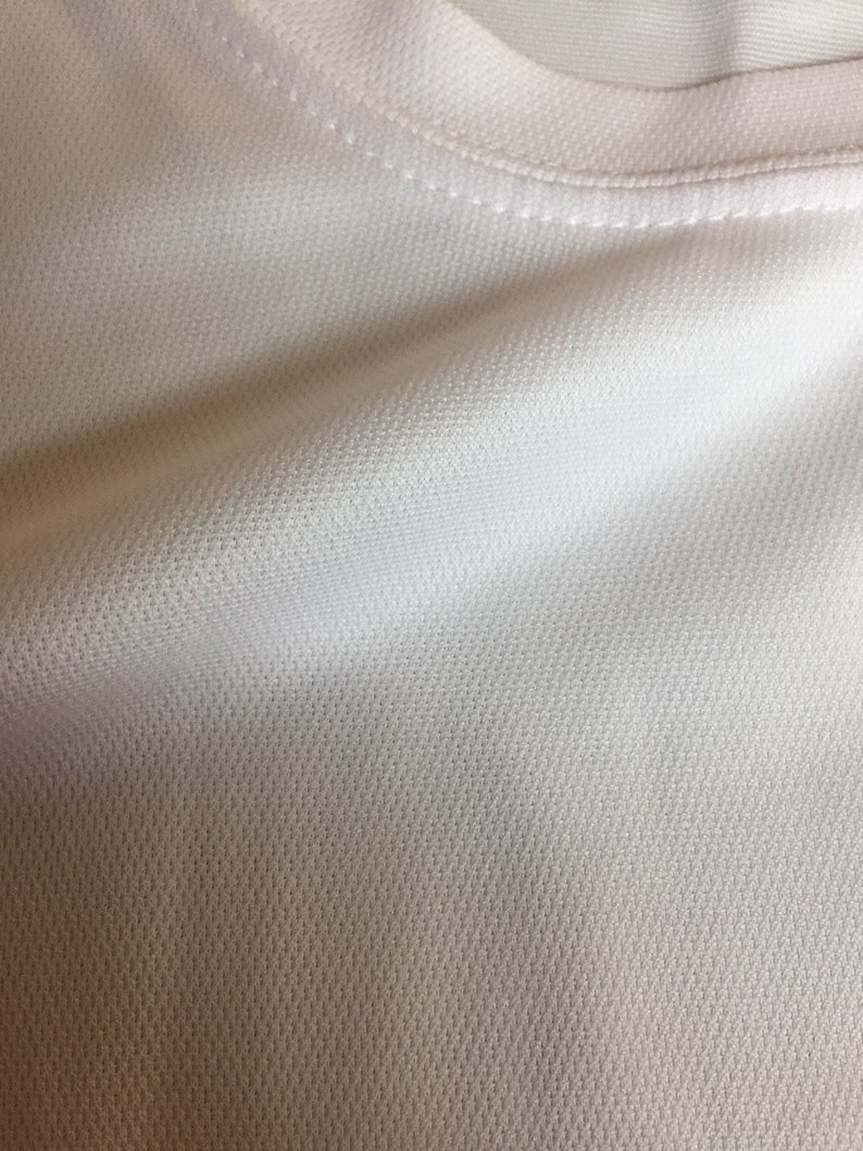 May include: Close-up of a white, textured fabric with a subtle sheen. The fabric has a fine, even weave and a smooth, soft feel.