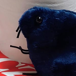 May include: A plush, dark blue creature with black antennae and small black eyes. The toy is positioned in front of a white object and a red surface with white text. The creature has a soft, fuzzy texture.