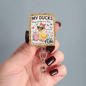 May include: A rectangular badge reel with a gold glitter border. The badge features a cartoon duck wearing sunglasses and a bow, holding a drink. Text on the badge reads "MY DUCKS May not be in a Row, But at least They are having FUN."