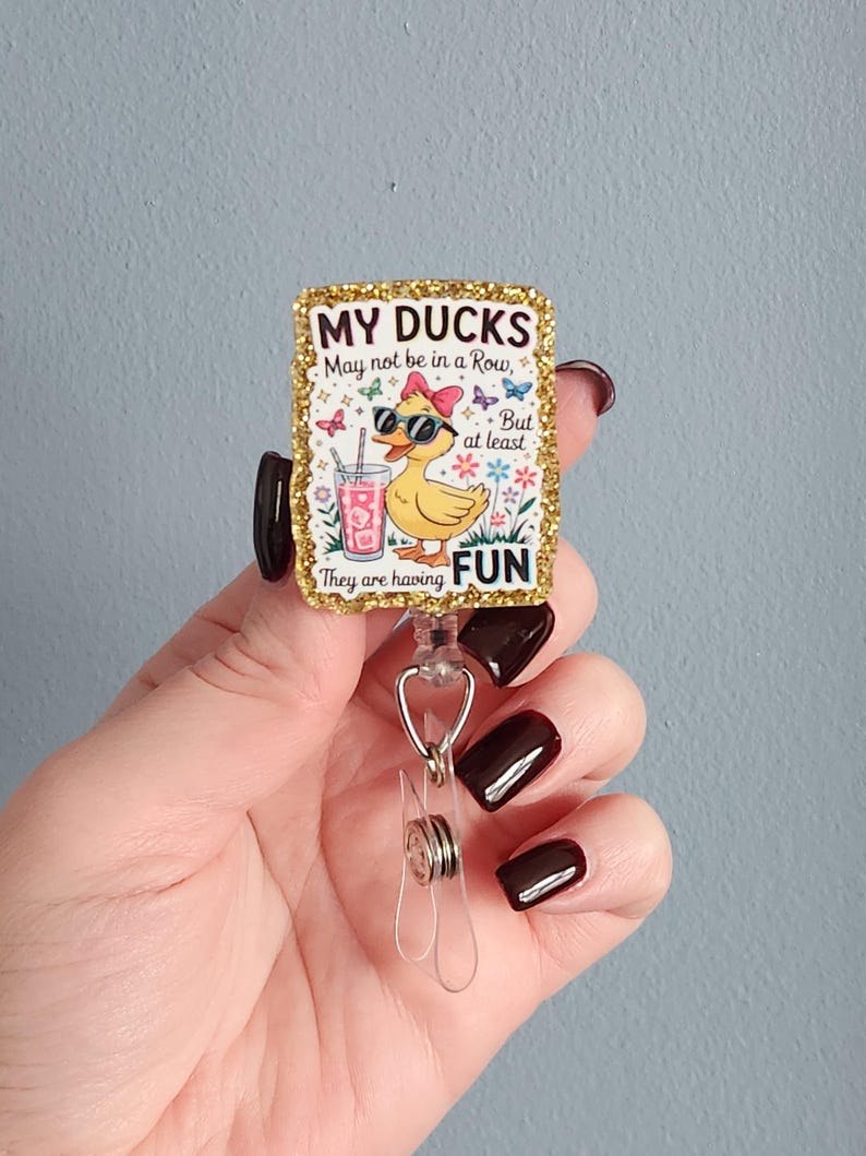 May include: A rectangular badge reel with a gold glitter border. The badge features a cartoon duck wearing sunglasses and a bow, holding a drink. Text on the badge reads "MY DUCKS May not be in a Row, But at least They are having FUN."