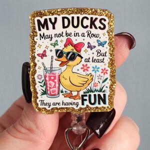 May include: A rectangular badge with a gold glitter border features a cartoon duck wearing sunglasses and a red bow. The text reads "MY DUCKS May not be in a Row, But at least They are having FUN."