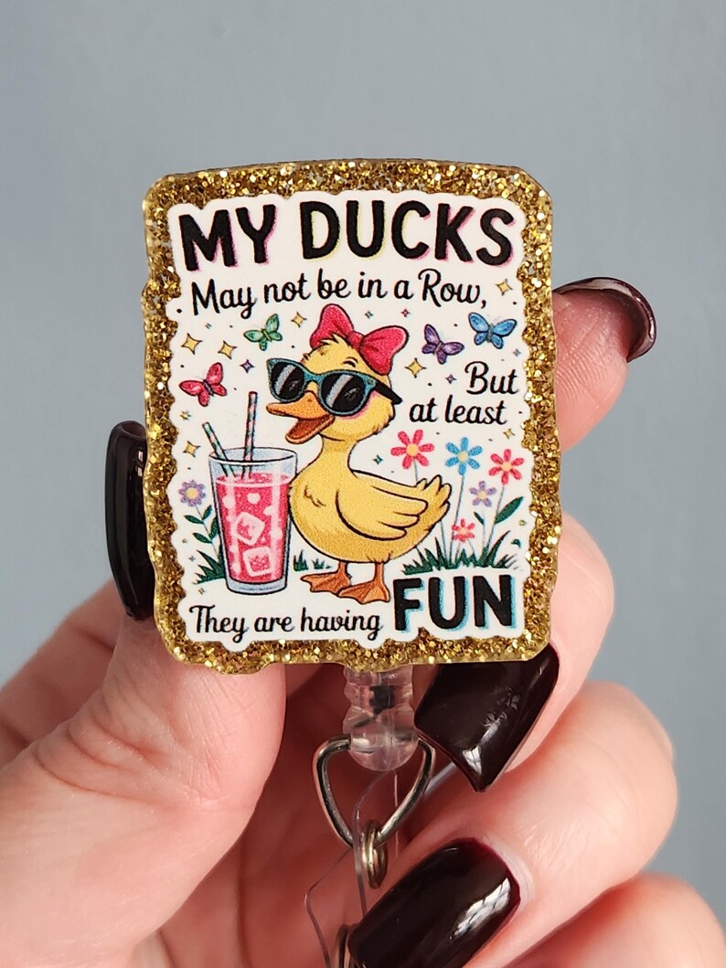 May include: A rectangular badge with a gold glitter border features a cartoon duck wearing sunglasses and a red bow. The text reads "MY DUCKS May not be in a Row, But at least They are having FUN."