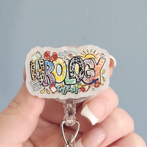 May include: A white and clear badge reel with the word "UROLOGY" in colorful, stylized letters. The design includes illustrations of kidneys, pills, and a sun. The reel has a clear plastic strap and a metal clip.