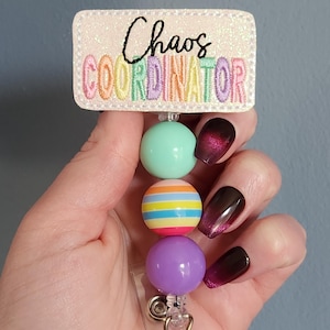 May include: A badge reel with the text "Chaos Coordinator" embroidered in black and rainbow colors on a glittery white background. The reel features three colorful beads: mint green, striped, and purple.