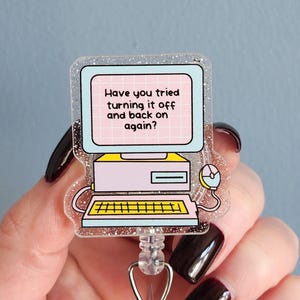 May include: A clear, glittery badge reel featuring a retro computer design. The computer screen displays the text "Have you tried turning it off and back on again?" in pink and white. The badge reel has a retractable cord and a clear plastic clip.