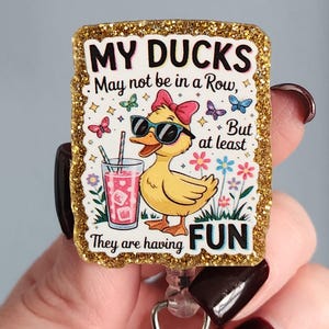 May include: A rectangular badge reel with a gold glitter border. The badge features a yellow duck wearing sunglasses and a pink bow, holding a drink. Text reads "MY DUCKS May not be in a Row, But at least They are having FUN."