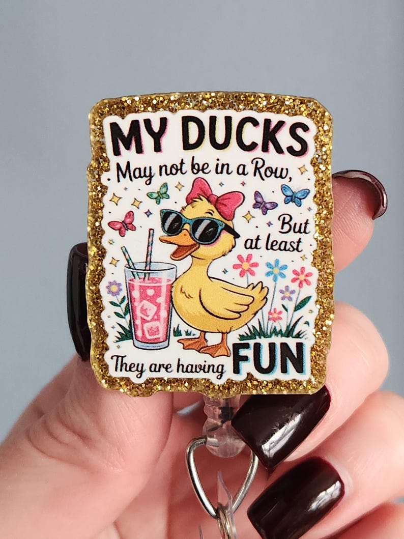 May include: A rectangular badge reel with a gold glitter border. The badge features a yellow duck wearing sunglasses and a pink bow, holding a drink. Text reads "MY DUCKS May not be in a Row, But at least They are having FUN."