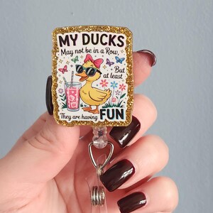 May include: A rectangular badge reel with a glittery gold border. The badge features a cartoon duck wearing sunglasses and a bow, holding a drink. Text reads "MY DUCKS May not be in a Row, But at least They are having FUN."