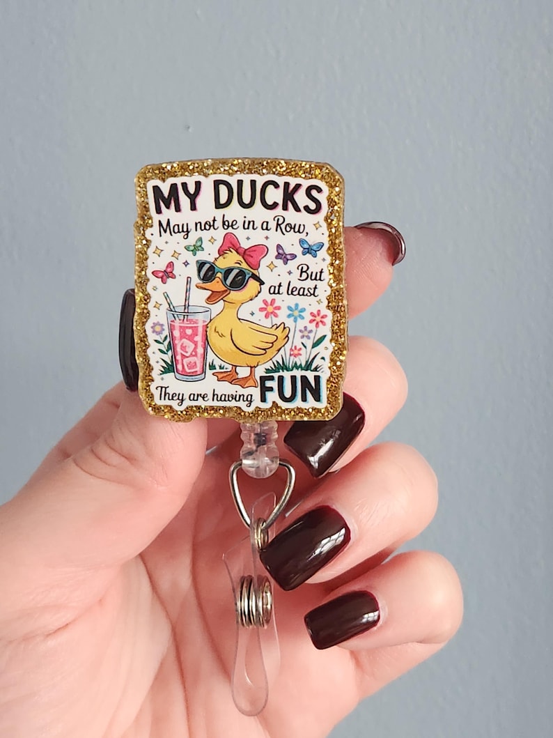 May include: A rectangular badge reel with a glittery gold border. The badge features a cartoon duck wearing sunglasses and a bow, holding a drink. Text reads "MY DUCKS May not be in a Row, But at least They are having FUN."