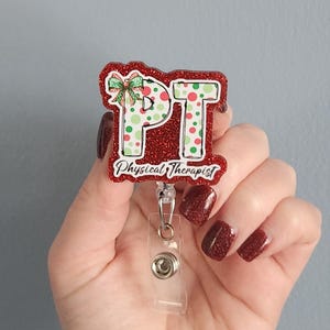May include: A red glitter badge reel with the letters "PT" in white, decorated with green and white polka dots and a green bow. The words "Physical Therapist" are written below the letters.