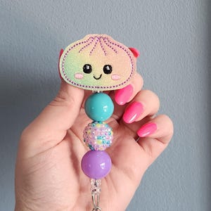 May include: A badge reel featuring a glittery, rainbow-colored embroidered dumpling character with a smiling face. The topper is attached to a vertical strand of three beads: one smooth turquoise, one white with multicolored rhinestones, and one smooth purple.