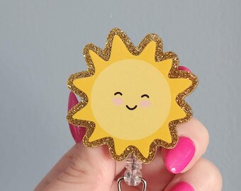 May include: A sun-shaped badge reel featuring a yellow face with a smiling expression and closed eyes. The sun has a thick, textured gold glitter border. A clear plastic strap and a metal clip are attached to the bottom of the sun-shaped piece.