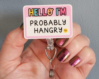 May include: A rectangular badge reel with the text "HELLO I'M PROBABLY HANGRY" in black, yellow, and pink. The badge reel has a clear plastic strap and a metal clip for attaching to clothing.